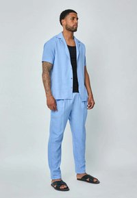 Chemise bleu clair à manches courtes avec un col décontracté, associée à un pantalon assorti et des sandales noires. Le tissu a une finition texturée.