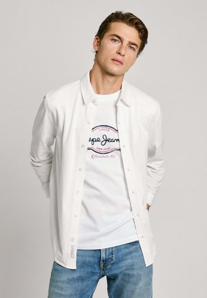 Weißes Hemd über einem weißen T-Shirt mit grafischem Druck in Blau und Rot, kombiniert mit hellblauen Jeans.