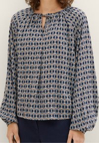 Blouse à manches longues présentant un motif géométrique en bleu marine, bleu clair et beige, avec un col en trou de serrure et un design d'épaule froncée.