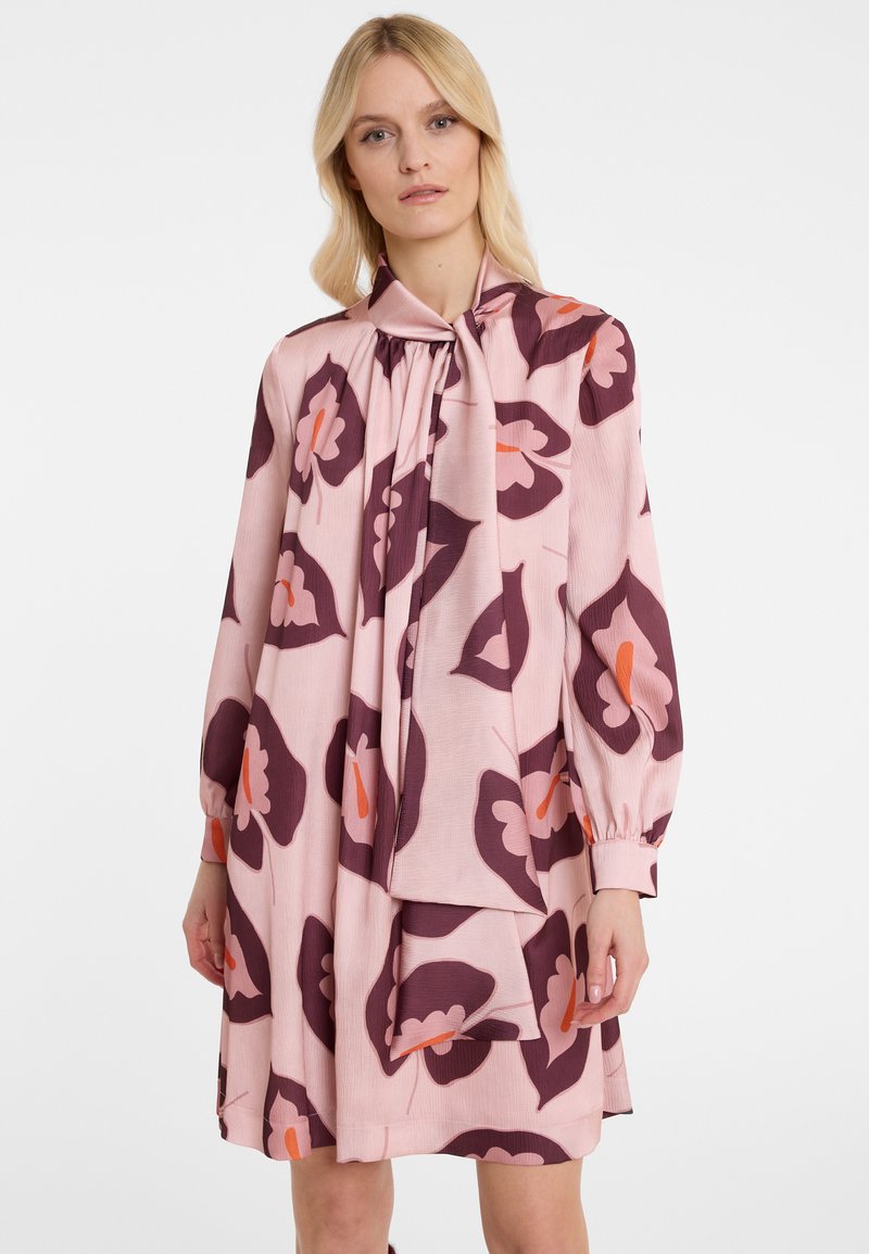 Frau in einem lockeren rosa Kleid mit großen dunkelvioletten und orangefarbenen Blumenmustern sowie einem gebundenen Kragen, stehend vor einem weißen Hintergrund.