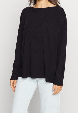 Femme portant un pull noir ample à manches longues avec un large col, associé à un jean bleu clair sur un fond uni.