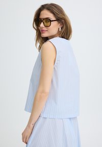 Femme aux cheveux ondulés portant de grandes lunettes de soleil teintées ambre, des boucles d'oreilles anneaux en argent, et un haut sans manches rayé bleu clair avec une jupe.