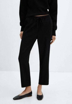 Frau trägt schwarze Hose mit Kordelzug, schwarzen Strickpullover und schwarze flache Schuhe und steht vor einem schlichten hellen Hintergrund.