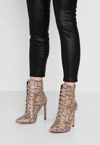 Snörda ankelstövlar i ett beige ormskinnsmönster, med spetsig tå och stilettklack, mot ett par svarta läderbyxor.