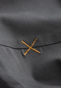 Zwarte stof met een gestructureerde weving, voorzien van een zichtbaar oranje gestikte "X"-detail op de naad, dat vakmanschap benadrukt.