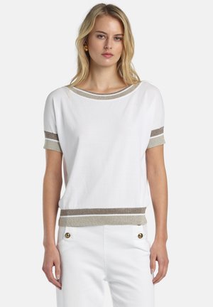 Donna che indossa una maglietta bianca a maniche corte con righe beige e marroni su collo, maniche e orlo, abbinata a pantaloni bianchi con bottoni dorati.