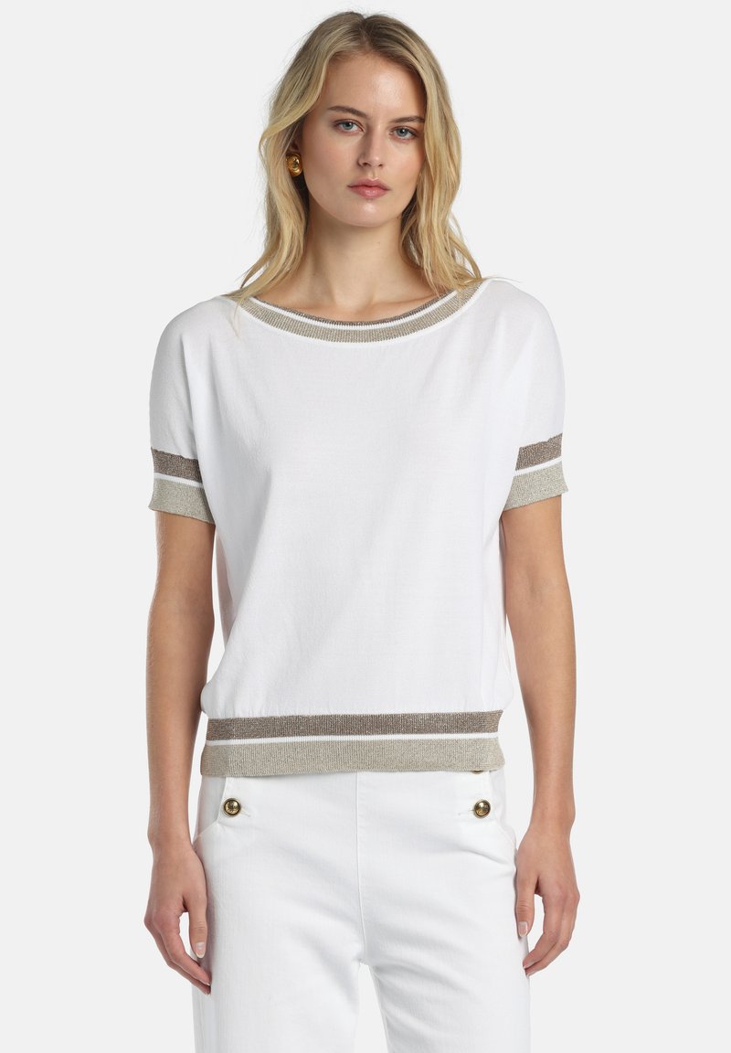 Sieviete valkā baltu īsroku kreklu ar bēšām un brūnām svītrām uz apkakles, piedurknēm un apakšmalas, kopā ar baltām biksēm ar zelta pogām.