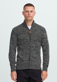 Hombre con cabello corto, vistiendo un suéter de punto gris con cremallera y pantalones negros, de pie contra un fondo claro y liso.