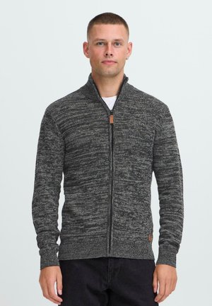 Hombre con cabello corto, vistiendo un suéter de punto gris con cremallera y pantalones negros, de pie contra un fondo claro y liso.