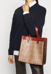 Brun mønstret totebag med røde læderdetaljer og solide håndtag, der har en rektangulær form og en glat overflade.