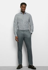 Hemd mit Knöpfen und blau-weißen Streifen, kombiniert mit grauer Anzughose und schwarzen Schuhen, stehend vor einem schlichten weißen Hintergrund.