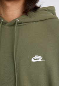 Ελιά πράσινη hoodie με κουκούλα και κορδόνι, που διαθέτει λευκό κεντημένο λογότυπο Nike στο στήθος. Λείο ύφασμα από βαμβάκι.
