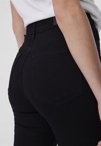 High-waisted black jeans featuring a smooth texture, five-pocket design, and subtle stitching details. Back view shows a fitted silhouette.
