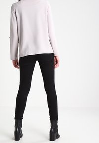 Haut gris clair à manches longues avec une coupe droite, associé à un jean skinny noir et des bottes noires à talons, vu de dos.
