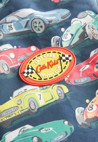 Tissu en coton coloré présentant divers voitures de dessin animé. Un patch en caoutchouc rouge et jaune indique "FAST CARS Cath Kids DRIVERS CLUB" avec des accents à damier.