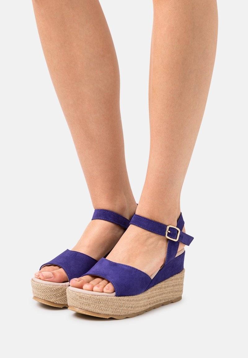 Pieds portant des sandales compensées en daim violet avec des brides chevilles et des fermetures à boucle sur une semelle plateforme tissée claire.
