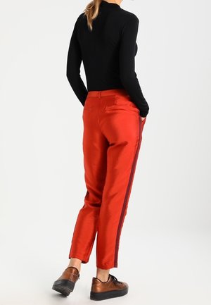 Pantalons en satin rouge avec des rayures sur les côtés, jambes fuselées et poches arrière, associés à un haut noir à manches longues et des mocassins marron.