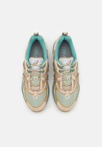 Scarpe da ginnastica beige e verde menta realizzate in materiali di rete e pelle, con un design curvo distintivo e un sistema di allacciatura elastica.