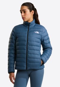 Woman with dark hair in a blue quilted jacket with front zipper and logo, standing with one hand in pocket, looking left.