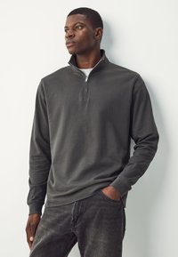 Felpa con cappuccio grigio scuro con collo a mezza zip, maniche lunghe, texture morbida e vestibilità comoda, abbinata a jeans scuri.