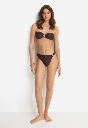 Woman stands barefoot wearing brown strapless bikini with gold ring details, against a plain white background.