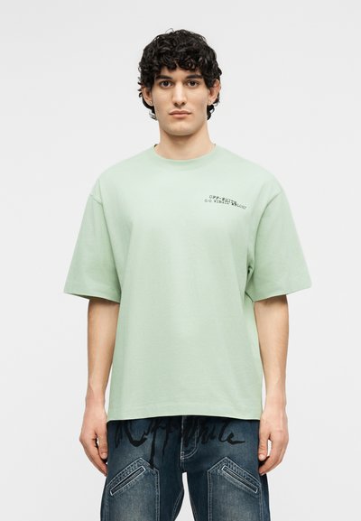 Jeune homme aux cheveux bouclés et foncés portant un t-shirt oversize vert pastel avec un texte noir sur la poitrine et un jean bleu foncé avec un imprimé graphique.