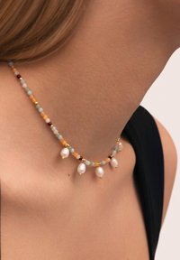 Collier perlé doré mettant en valeur des perles bleu clair, ambre et bordeaux avec des accents de coquillage blanc pendants, sur une surface de peau lisse.