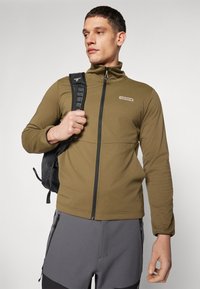 Olive green zip-up jacket with a high collar, made of soft fabric. Features a zippered pocket and is paired with gray shorts and a black backpack.