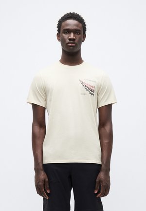 Hellbeiges T-Shirt mit kurzen Ärmeln aus weichem Material, mit einem bunten, gemusterten Grafikdesign auf der linken Brustpartie.