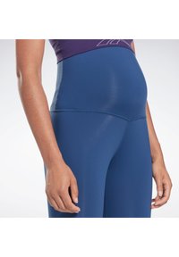 Leggings azuis de cintura alta, com tecido elástico, textura suave e um ajuste contornado. Acentuados com uma faixa roxa na cintura e detalhes nas costuras.