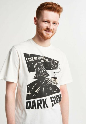 Rothaariger Mann trägt weißes T-Shirt mit Darth Vader, der eine Kaffee-Tasse mit der Aufschrift "#1 Dad" hält, und dem Text "Ich mag meinen Kaffee auf der dunklen Seite."