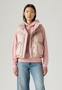 Gevoerde, lichtroze vest met een ritssluiting aan de voorkant en zijzakken, gelaagd over een bijpassende roze hoodie. Deze outfit is gestyled met denim jeans.