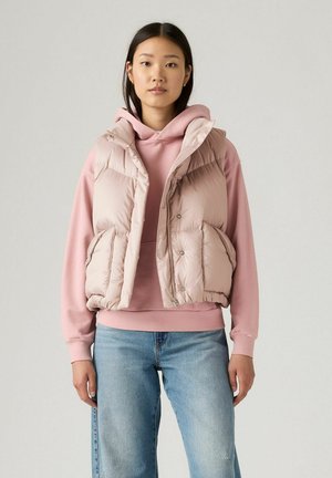 Gevoerde, lichtroze vest met een ritssluiting aan de voorkant en zijzakken, gelaagd over een bijpassende roze hoodie. Deze outfit is gestyled met denim jeans.