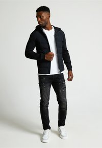 Crna puffer jakna sa prilagođenim dizajnom, nosi se preko bijele majice. Sivi poderani traperice i bijele tenisice upotpunjuju izgled.