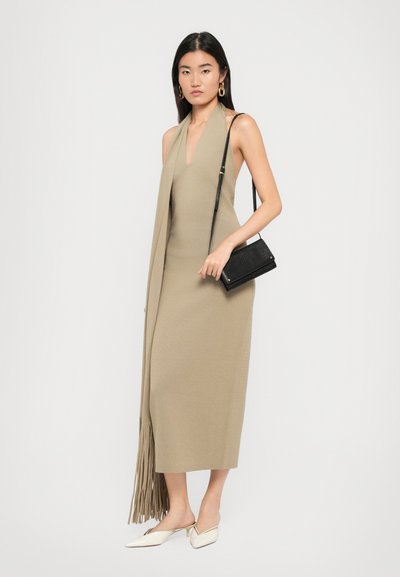 Woman in beige sleeveless midi dress with long fringe scarf, white mules, gold hoop earrings, and black shoulder bag on white background.