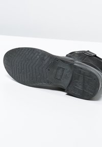 Bota de cuero negra con una suela de goma plana y texturizada. Presenta un detalle de hebilla en el lateral y un logotipo de la marca en relieve en la suela.