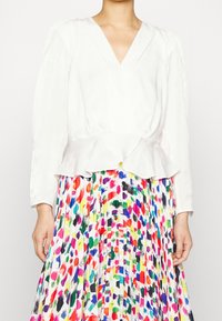 Blusa blanca con peplum, de manga larga y escote en V, combinada con una falda plisada colorida que presenta un patrón de manchas de pintura en varios tonos.