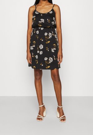 Vrouw draagt een zwarte mouwloze bloemenjurk met witte en gele bloemen en witte sandalen met hak, staand tegen een effen achtergrond.