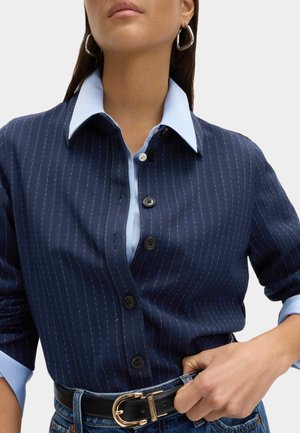 Kvinde iført marineblå skjorte med lyseblå krave og manchetter med nålestriber, stoppet ned i jeans med sort bælte.
