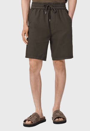 Mann trägt dunkelbraune elastische Shorts mit Gummibund und braune gewebte Slip-on-Sandalen, steht mit den Händen in den Taschen.