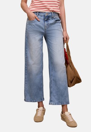 Kobieta w jasnoniebieskich szerokich spodniach dżinsowych o długości za kostkę, beżowych tenisówkach, czerwono-białej koszuli w paski, trzyma brązową torbę typu tote z czerwoną apaszką w środku.