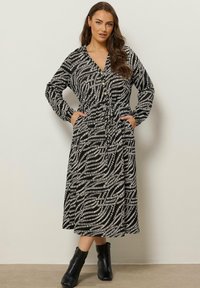 Kobieta w czarno-białej abstrakcyjnej sukience midi z długim rękawem i czarnych botkach za kostkę, stojąca z rękami w kieszeniach.
