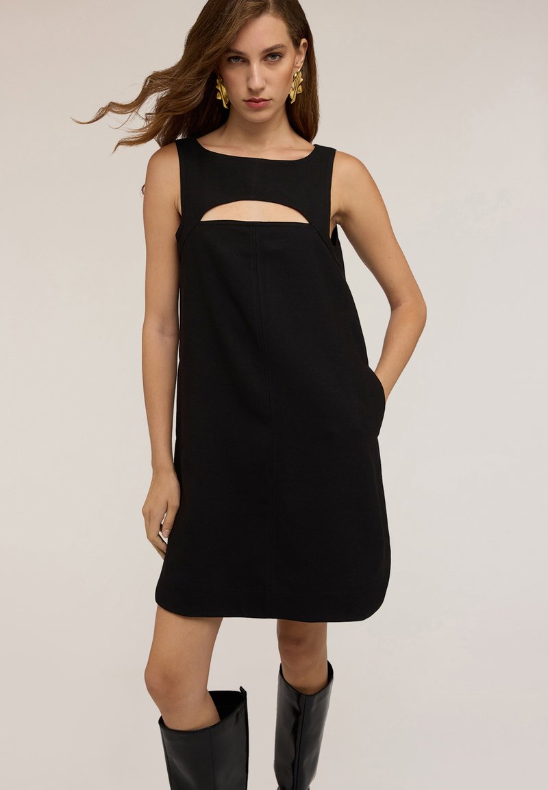 Vestido negro sin mangas con un detalle recortado en el pecho, silueta recta, bolsillos laterales y una textura suave.