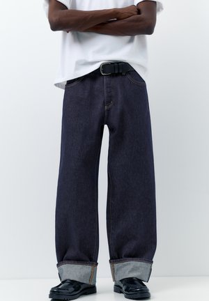 Személy, aki széles szárú sötétkék farmert visel felhajtott szegéllyel, fehér pólót, fekete övet és fekete fényes loafer cipőt, karját keresztbe téve áll.