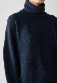 Pulover de culoare bleumarin cu guler înalt, realizat dintr-un material tricotat moale. Prezintă o textură canalată și o croială lejeră, cu umeri puțin căzuți.