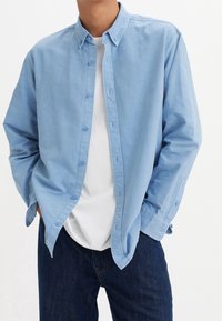 Chemise à boutons bleu clair en coton, avec un col classique, des manches longues et une poche poitrine unique, portée par-dessus un t-shirt blanc.