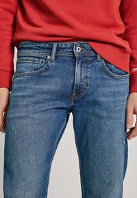 Jeans de mezclilla azul claro con corte recto, diseño de cinco bolsillos y detalles de costura visibles. Llevados con un sudadera roja.