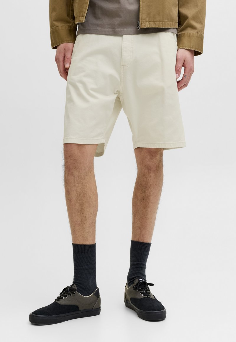 Person iført cremefarvede knælange shorts, sorte sokker og sorte og olivengrønne sneakers, stående mod en lys baggrund.