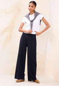 Valge lühikeste varrukatega T-särk, mida on kombineeritud laia säärtega tumesiniste pükstega; hall kampsun on seotud õlgade ümber; modell seisab heleda tekstuuriga tausta ees.