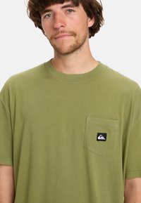 Olive green cotton t-shirt with a crew neck, short sleeves, and a left chest pocket featuring a black and white logo patch.